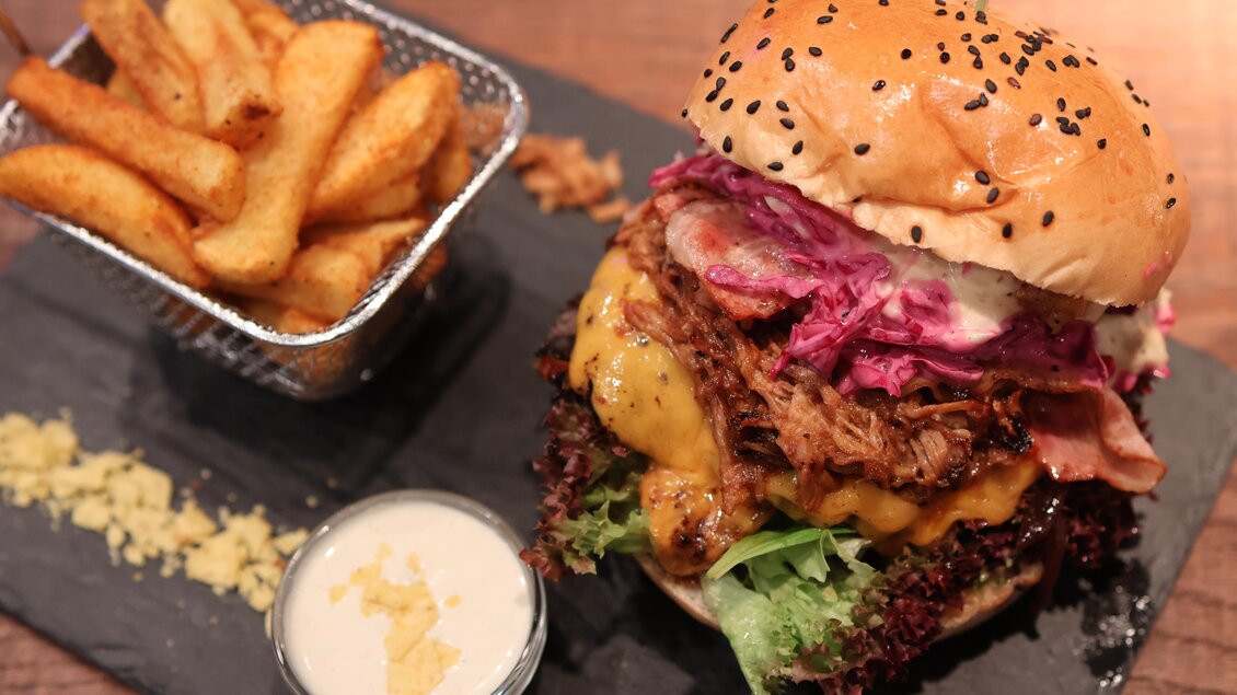Ein großer Burger mit geschmolzenem Käse, rotem Kraut und frischem Salat. Dazu gibt es knusprige Kartoffelspalten und eine kleine Schale mit Dip. | © kAlps Foodbar
