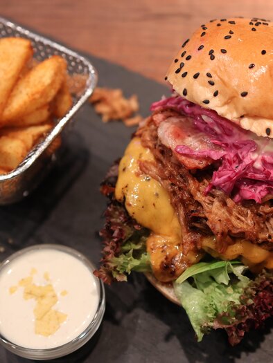 A large burger with melted cheese, red cabbage, and fresh lettuce. Served with crispy potato wedges and a small bowl of dip. | © kAlps Foodbar