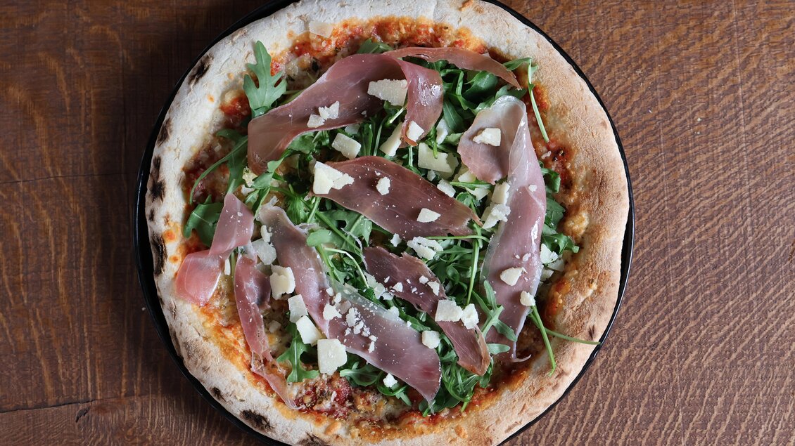 Eine köstliche Pizza mit frischem Rucola, Parmaschinken und Käse. Der knusprige Rand und der bunte Belag verleihen ihr einen ansprechenden Look. | © kAlps Foodbar