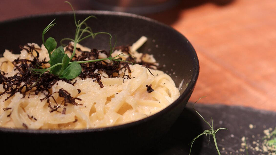 Ein Teller Pasta in einer schwarzen Schüssel, garniert mit Kräutern und schwarzem Trüffel. Die Schüssel steht auf einem Holz- und Steinsockel, umgeben von frischen Kräutern. | © kAlps Foodbar
