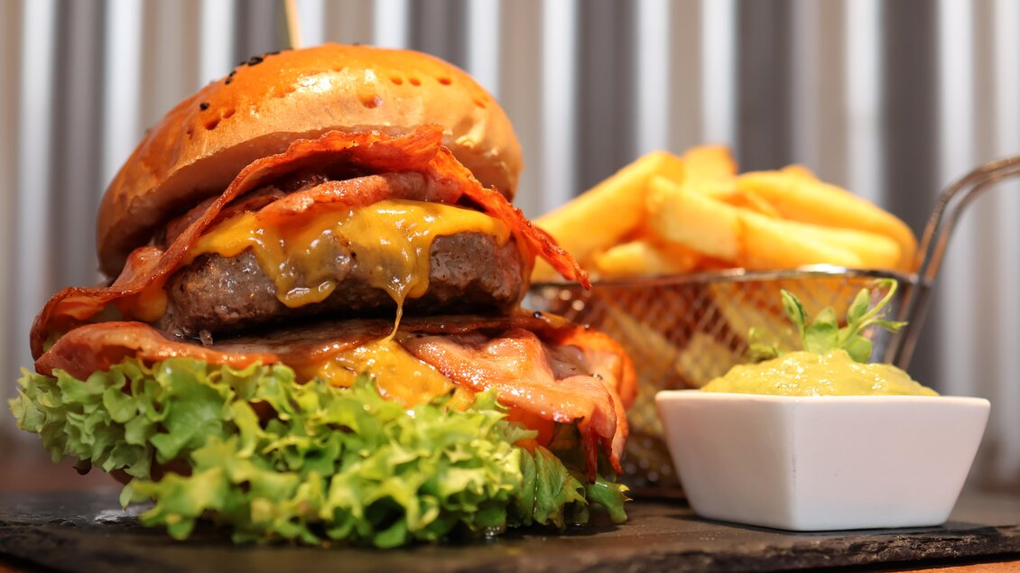 Ein saftiger Burger mit Käse, Speck und frischem Salat auf einem schwarzen Teller. Neben dem Burger befinden sich Pommes frites und eine kleine Schüssel mit einer grünen Sauce. | © kAlps Foodbar 