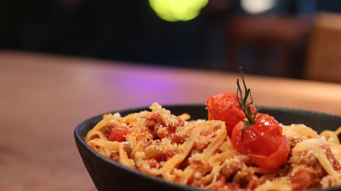 Ein köstlicher Teller Spaghetti mit Hackfleischsauce, garniert mit Kirschtomaten. Der Teller steht auf einem dunklen Untergrund und ist von einer gemütlichen Atmosphäre umgeben. | © kAlps Foodbar