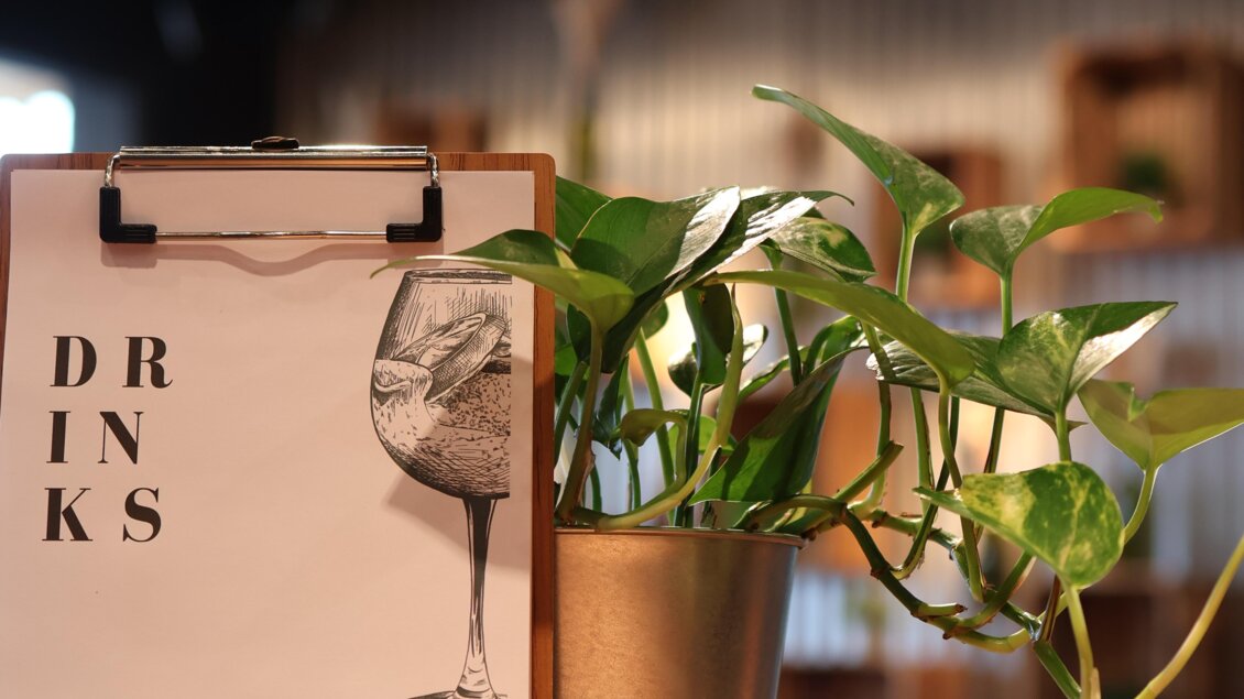Ein Getränkemenü steht auf einem Tisch, neben einer grünen Pflanze in einem silbernen Topf. Die Umgebung ist gemütlich und modern gestaltet. | © kAlps Foodbar