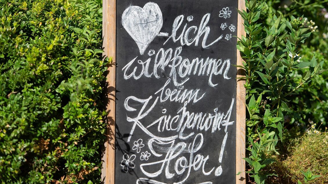 Eine Schiefertafel mit der handgeschriebenen Begrüßung „Willkommen beim Kuchenpunkt Hofer!“ umgeben von grünen Pflanzen. Herzen und Blumen verleihen der Tafel einen freundlichen Touch. | © kh Kirchenwirt Hofer