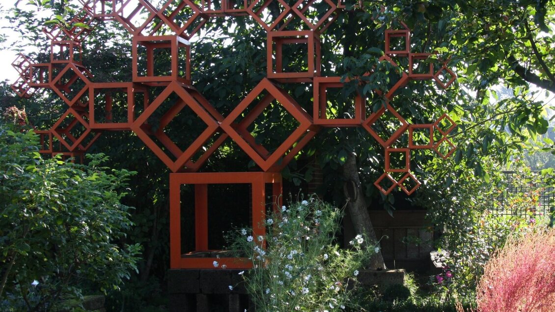 Orange Metallskulptur aus Quadraten im kunstGarten Graz, umgeben von Pflanzen | © kunstGarten