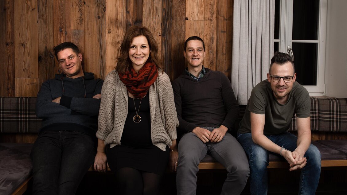 Eine Gruppe von vier Personen sitzt auf einer Bank vor einer Holzwand. Sie lächeln und scheinen eine entspannte Zeit zusammen zu haben. | © Nadine Geuter