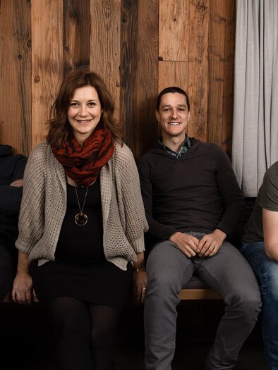 Eine Gruppe von vier Personen sitzt auf einer Bank vor einer Holzwand. Sie lächeln und scheinen eine entspannte Zeit zusammen zu haben. | © Nadine Geuter