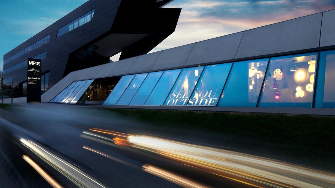 Ein modernes Gebäude mit großen Fenstern, durch die eine kreative Ausstellung zu sehen ist. Die Fassade ist stilvoll und futuristisch gestaltet. | © nullneun - Gerald Liebminger