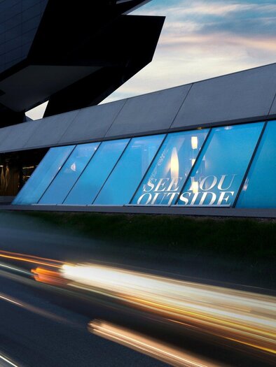 Ein modernes Gebäude mit großen Fenstern, durch die eine kreative Ausstellung zu sehen ist. Die Fassade ist stilvoll und futuristisch gestaltet. | © nullneun - Gerald Liebminger