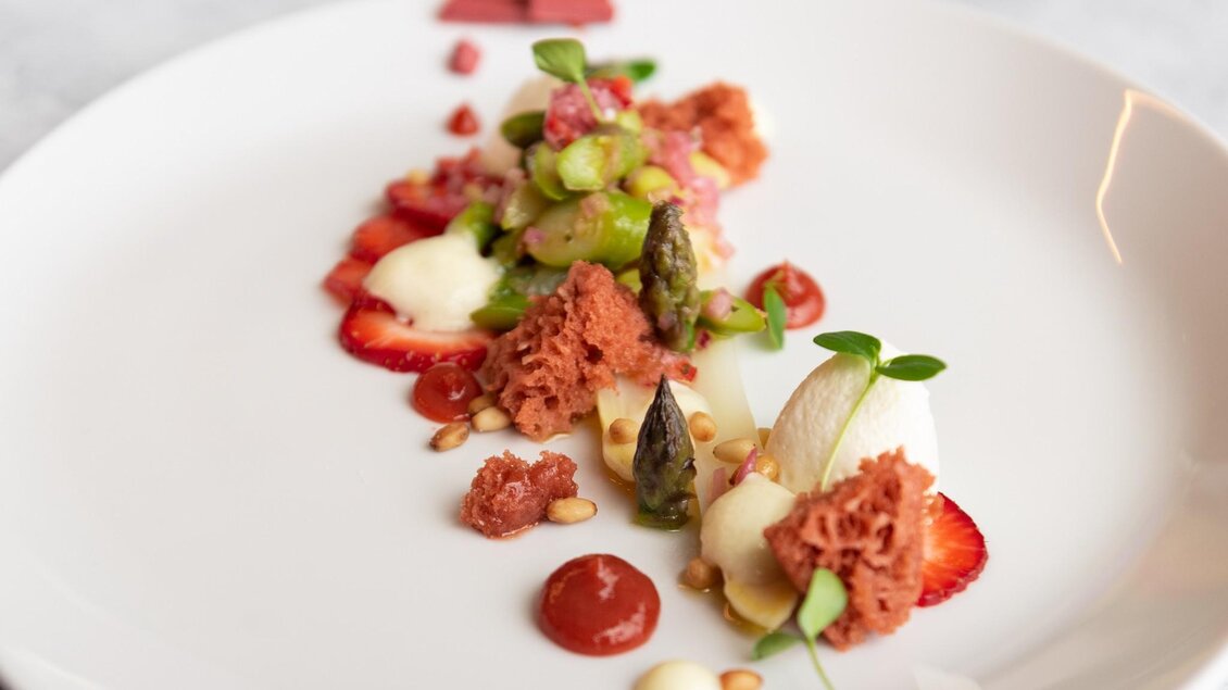 Modern angerichtetes Gericht mit Spargel, Erdbeeren, Creme, Soßentupfen, Pinienkernen und luftigem Biskuit auf weißem Teller. | © nullneun