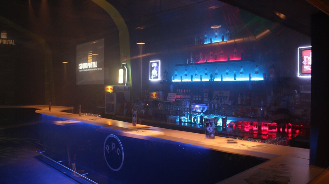 Eine stilvolle Bar mit einer beleuchteten Theke und bunten Drinks. Im Hintergrund sind leuchtende Flaschen und digitale Displays zu sehen. | © ppc