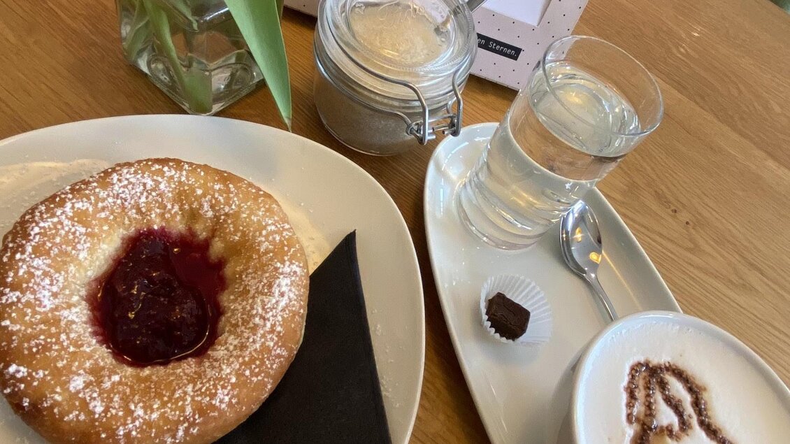 Ein köstlicher Gebäck mit Marmelade, serviert auf einem Teller mit schwarzem Papier. Dazu gibt es eine Tasse Kaffee und ein Glas Wasser, sowie eine Vase mit Tulpen. | © pur Styria - der Dorfladen