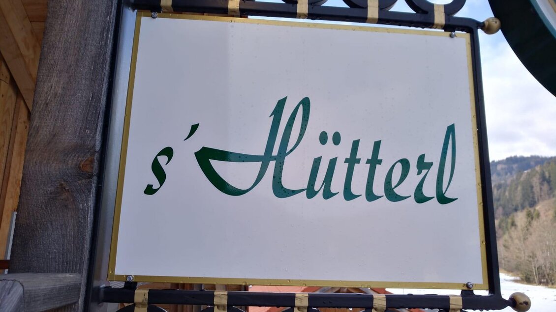 Ein Schild mit der Aufschrift „s'Hütterl“. Es befindet sich in einer ländlichen Umgebung mit schöner Landschaft. | © Jürgen Strohmeyer