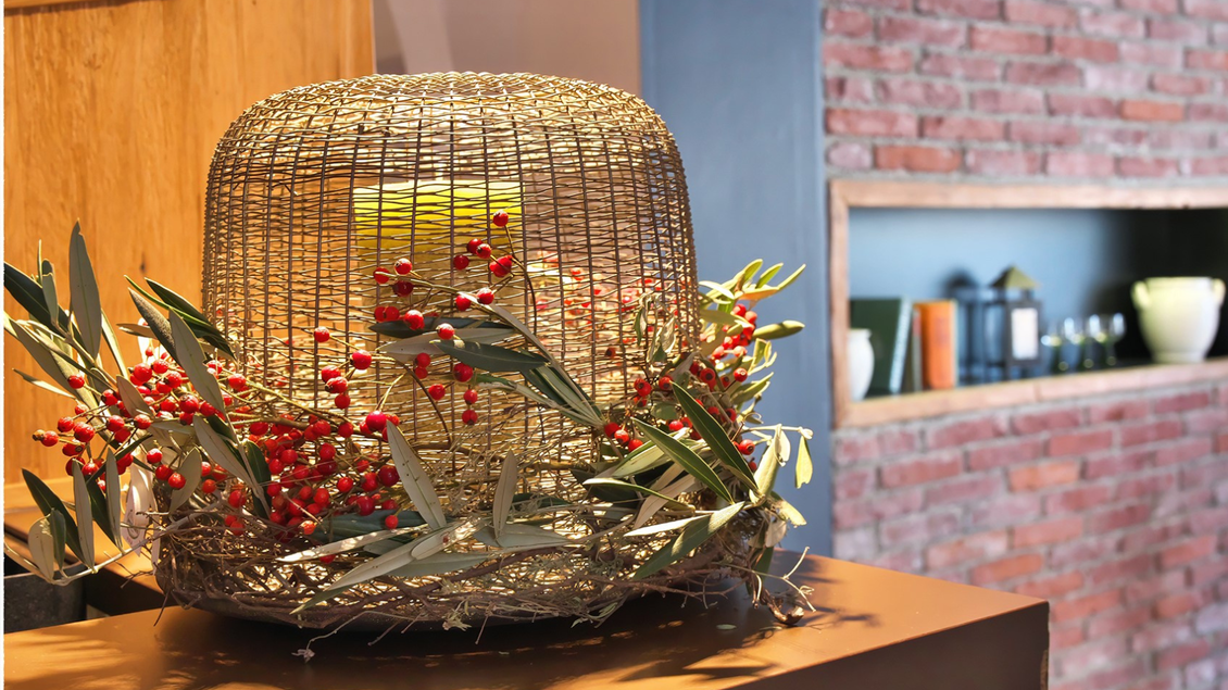 Ein dekorativer Korb mit roten Beeren und grünen Blättern steht auf einem Tisch. Im Hintergrund sind Regale mit verschiedenen Gegenständen sichtbar. | © event-fotograf.at