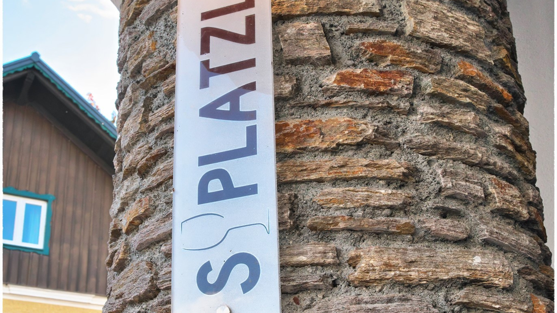 Ein Schild mit der Aufschrift "S PLATZL" an einem steinernen Pfeiler. Im Hintergrund ist ein traditionelles Gebäude zu sehen. | © event-fotograf.at
