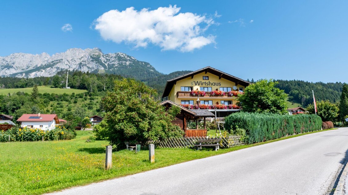 Ein malerisches Gasthaus mit bunten Blumenkästen und Bergen im Hintergrund. Die Umgebung ist grün und sonnig, ideal für einen entspannten Aufenthalt. | © s'Wirtshaus