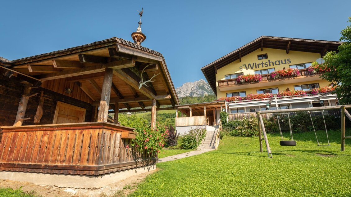 Ein traditionelles Wirtshaus mit einem Holzgebäude und einem gepflegten Garten. Im Hintergrund sind blühende Balkone und ein klarer Himmel zu sehen. | © s'Wirtshaus