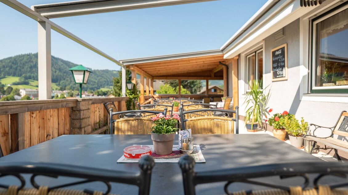 Eine Terrasse mit Tischen und Stühlen, umgeben von Pflanzen. Der Blick zeigt eine Landschaft mit Hügeln und klarem Himmel. | © s'Wirtshaus