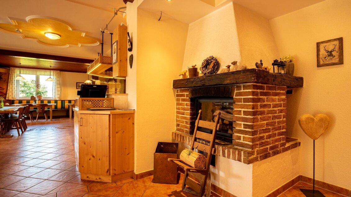 Ein gemütlicher Wohnraum mit einem Kamin aus Backstein. Der Raum ist hell und einladend, mit einem Holzstuhl und einer offenen Küche im Hintergrund. | © s'Wirtshaus