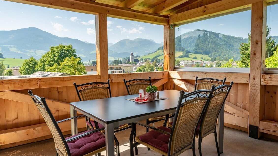 Ein gemütlicher Essbereich auf einer Veranda mit Holzverkleidung und Blick auf die Berge. Der Tisch ist für sechs Personen gedeckt und es gibt einen kleinen Pflanze in der Mitte. | © s'Wirtshaus