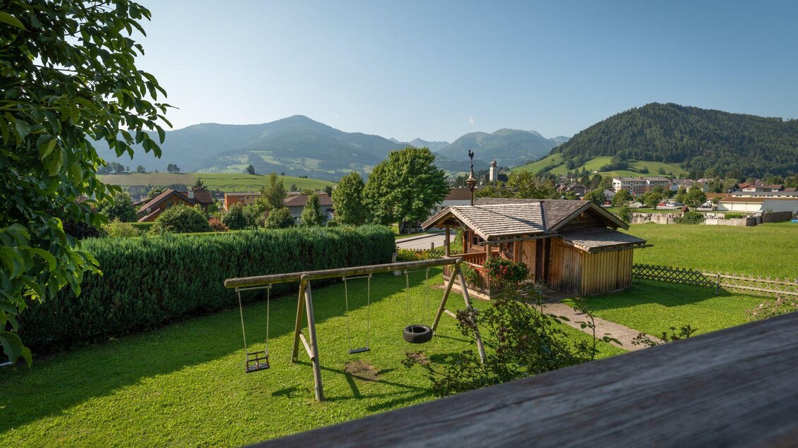Ein malerischer Ausblick auf eine ländliche Umgebung mit einem Holzhaus und einem Spielplatz. Im Hintergrund erheben sich sanfte Berge unter einem klaren Himmel. | © s'Wirtshaus