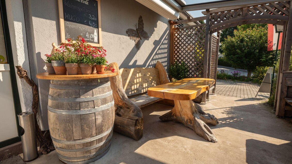 Eine gemütliche Terrasse mit einem Holztisch und einer Sitzbank. Auf einem Fäßchen stehen blühende Pflanzen. | © s'Wirtshaus