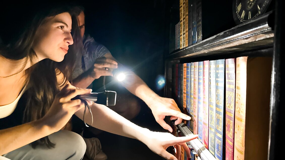 Eine Frau und ein Mann untersuchen eine Bücherregal mit Taschenlampen. Sie scheinen nach etwas Bestimmtem in den alten Büchern zu suchen. | © Apolla GmbH