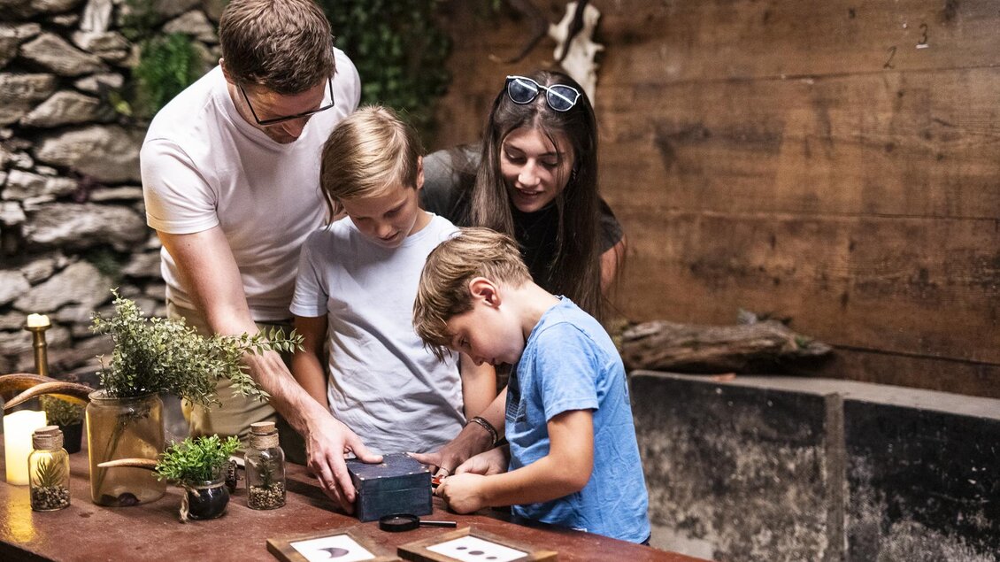 Eine Familie steht um einen Tisch und untersucht einen kleinen Gegenstand. Im Hintergrund sind steinerne Wände und Holzdetails sichtbar. | © Apolla GmbH