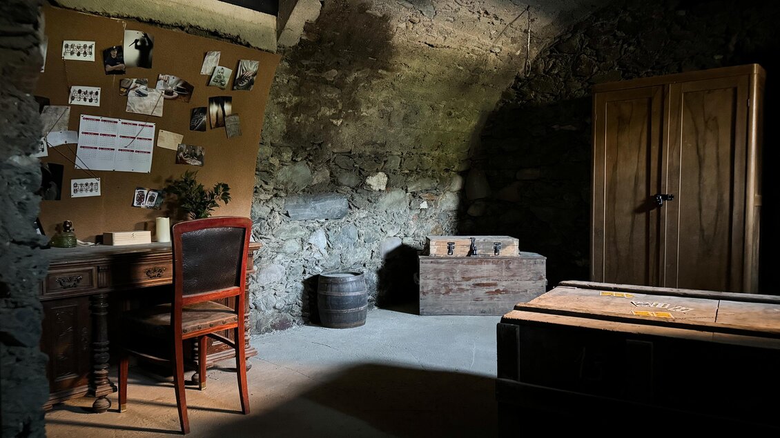 Ein dunkles, nostalgisches Zimmer mit steinernen Wänden. Ein Schreibtisch mit einem Stuhl und einigen Kisten ist im Vordergrund zu sehen. | © Apolla GmbH