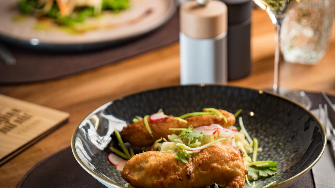 Nahaufnahme eines gedeckten Tisches mit einem Teller leckerer, goldbraun gebackener Speisen, garniert mit frischen Kräutern und Gemüse. Im Hintergrund steht ein Glas Weißwein und ein weiteres Tellergericht. | © Vagiss mi nit