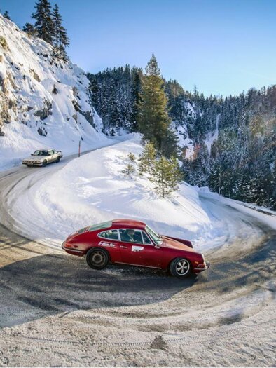 Two cars are driving in a snow-covered mountain landscape. The sun is shining through the trees, making the scene appear radiant. | © Markus Tobisch
