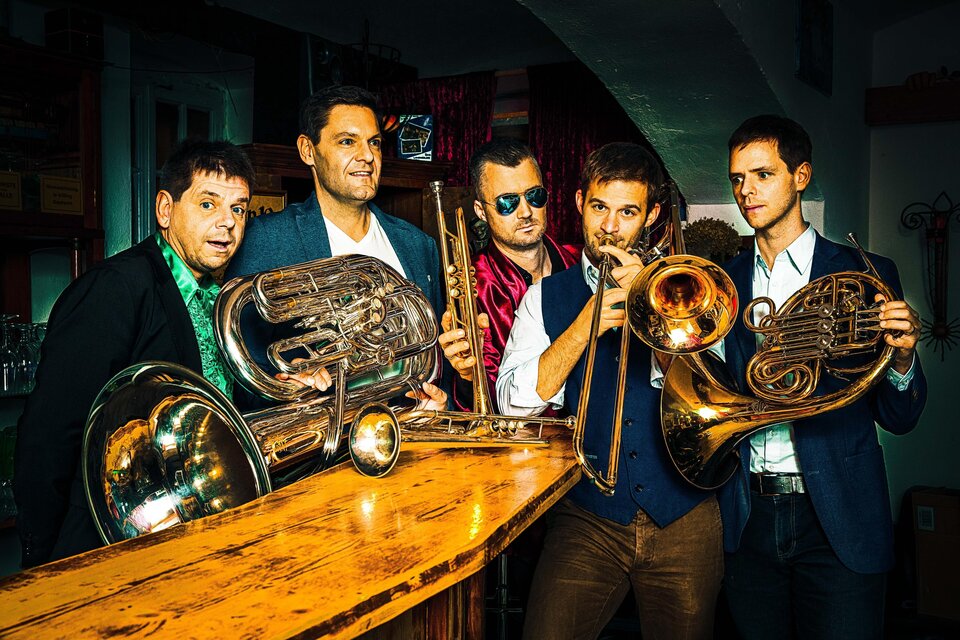 Eine Gruppe von fünf Musikern steht an einer Bar und hält verschiedene Blasinstrumente. Sie wirken lässig und präsentieren ein fröhliches Ambiente. | © Magic Brass