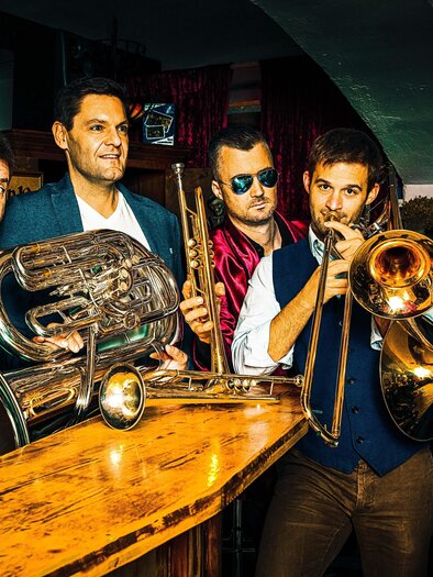 Eine Gruppe von fünf Musikern steht an einer Bar und hält verschiedene Blasinstrumente. Sie wirken lässig und präsentieren ein fröhliches Ambiente. | © Magic Brass