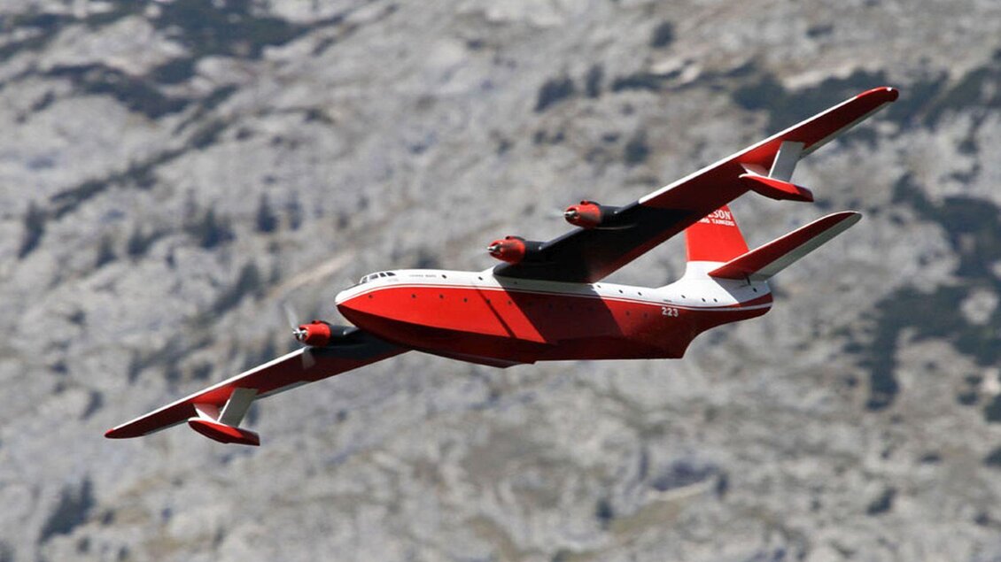 Ein großes Flugzeug mit rotem und weißem Design fliegt hoch über einer bergigen Landschaft. Die Tragflächen sind weit gespannt und die Landschaft im Hintergrund zeigt felsige Strukturen. | © Ian Walter