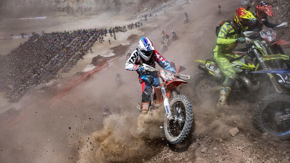 Motorradfahrer kämpfen sich durch staubige, felsige Gelände in einem aufregenden Wettkampf. Im Hintergrund sind viele Zuschauer zu sehen, die das Rennen verfolgen. | © Erzbergrodeo GmbH