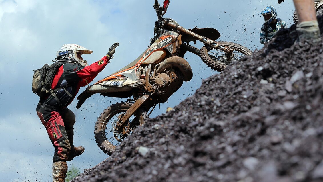 Ein Motocross-Fahrer fährt einen steilen Hang hinauf. Die Umgebung ist steinig und staubig, mit einem klaren Himmel im Hintergrund. | © Tiqa