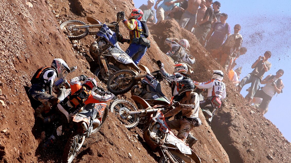 Eine aufregende Motocross-Veranstaltung auf einem steilen Hang. Mehrere Fahrer kämpfen mit ihren Motorrädern, während Zuschauer zuschauen. | © Tiqa
