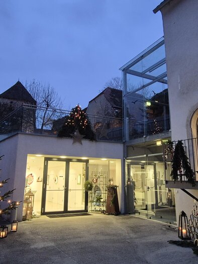 Ein einladender Eingang zu einem Gebäude mit Weihnachtsbeleuchtung und einem Christbaum. Der Himmel ist dämmerig und verleiht der Szenerie eine festliche Atmosphäre. | © Oststeiermark Tourismus