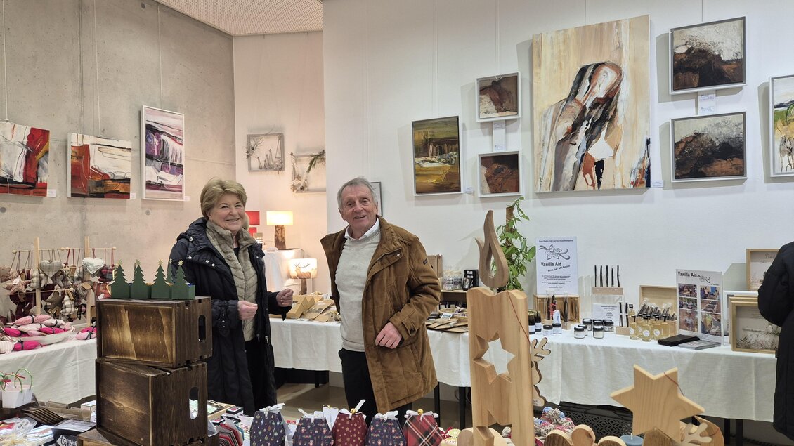 Ein Verkaufsstand mit Holzfiguren und handgefertigten Produkten. Zwei Personen stehen lächelnd im Raum mit Kunstwerken an der Wand. | © Oststeiermark Tourismus