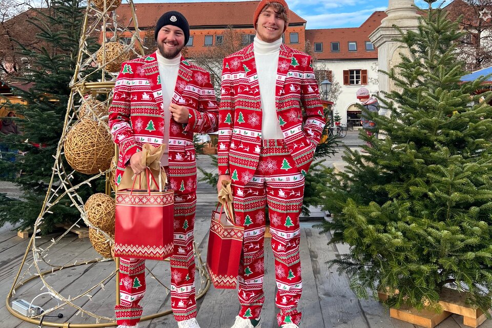 Zwei Männer in festlichen roten Anzügen mit Weihnachtsmotiv stehen auf einem Weihnachtsmarkt. Im Hintergrund sind geschmückte Bäume und eine festliche Dekoration zu sehen. | © Stadtmarketing
