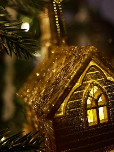 A golden Christmas tree ornament in the shape of a house with a glowing window. In the background, decorative lights shimmer. | © Pixabay.com