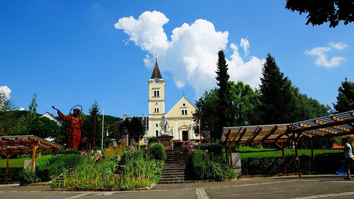 Eine malerische Kirche vor einem blauen Himmel mit weißen Wolken. Im Vordergrund sind grüne Pflanzen und eine ansprechende Parklandschaft zu sehen. | © TV Thermen- & Vulkanland