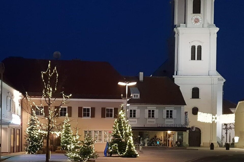 Ein schöner Platz bei Nacht mit festlich dekorierten Weihnachtsbäumen. Im Hintergrund steht ein markantes Gebäude mit einem Turm und einem Ziffernblatt. | © Stadtgemeinde Fehring