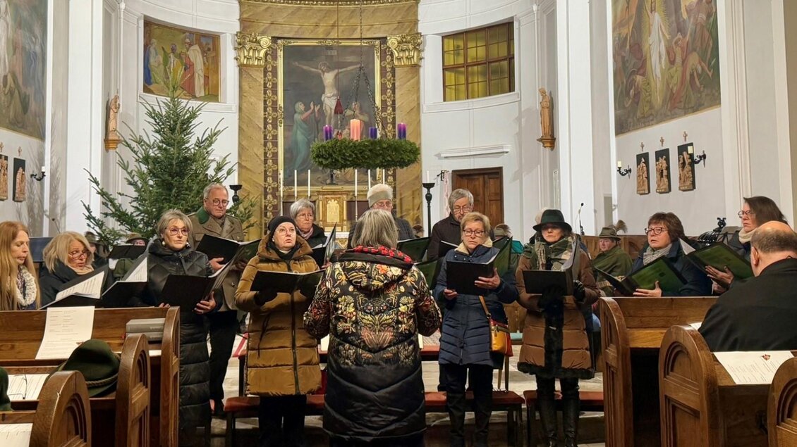 Ein Chor singt in einer Kirche, umgeben von festlicher Dekoration. Im Hintergrund steht ein Adventskranz und Weihnachtsbäume. | © Franz-Peter Stadler