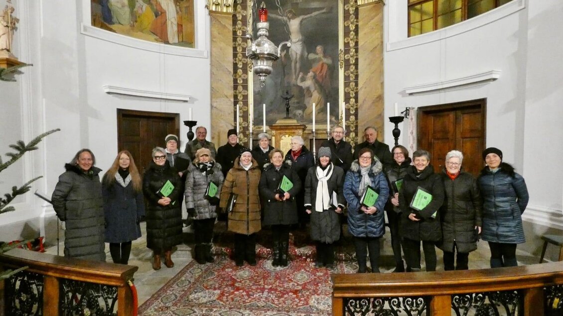 Eine Gruppe von Menschen steht in einer Kirche, die in warmen Farbtönen dekoriert ist. Sie halten Notenblätter in den Händen und scheinen bereit zu sein, zu singen. | © Hans Platzer