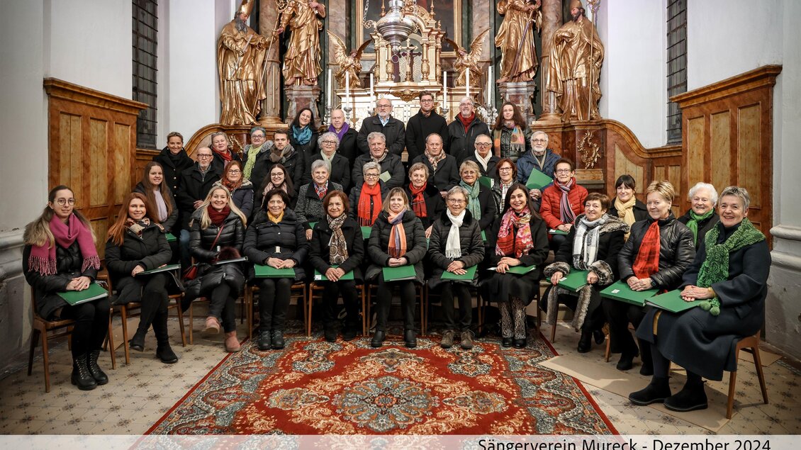Eine Gruppe von Sängern posiert in einer Kirche vor einem prächtigen Altar. Sie tragen schwarze Kleidung und halten grüne Blätter in den Händen. | © Fot de Monte