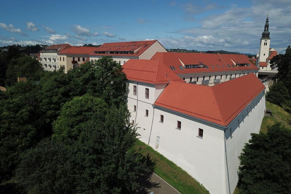 Ein historisches Gebäude mit rotem Dach und weißen Wänden. Im Hintergrund sieht man einen Kirchturm und viel Grün um das Gebäude herum. | © Austro-Pop-Zeitreise