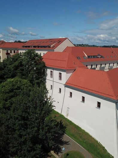 Ein historisches Gebäude mit rotem Dach und weißen Wänden. Im Hintergrund sieht man einen Kirchturm und viel Grün um das Gebäude herum. | © Austro-Pop-Zeitreise