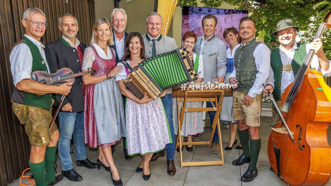 Das Team hinter dem Steiermark-Event 'Aufsteirern' posiert gemeinsam in traditioneller Tracht. Mit Instrumenten wie Akkordeon, Geige und Kontrabass in den Händen, stehen sie vor einem Hintergrund mit dem Hashtag '#Aufsteiern'. Sie strahlen eine freundliche und einladende Atmosphäre aus und repräsentieren die regionale Kultur und Musiktradition. | © Ivents Kulturagentur-Erwin Scheriau