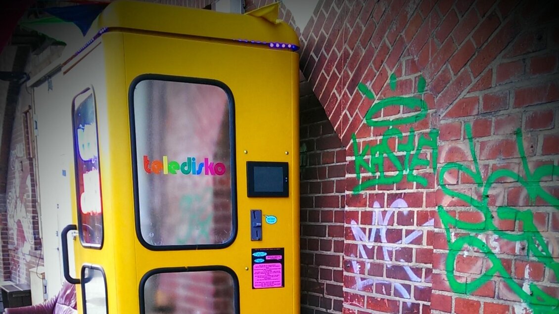 Ein gelbes Telefonhäuschen steht an einer Ziegelwand mit Graffiti. Im Hintergrund sind farbige Bänder zu sehen. | © teledisko